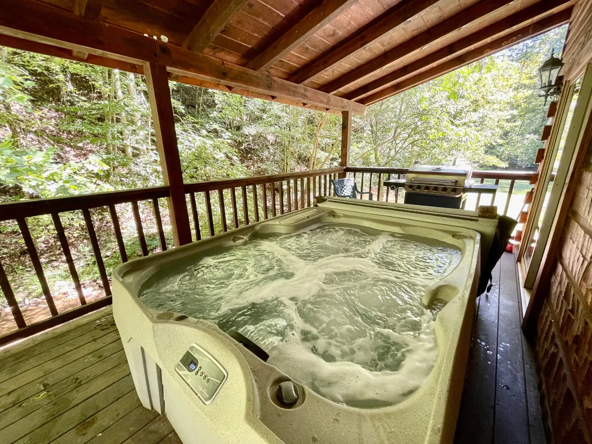 A tranquil scene featuring a bubbling hot tub on a wooden deck, surrounded by lush greenery. A propane grill sits nearby, while the deck overlooks a grassy yard, inviting guests to relax, cook outdoors, or soak in the peaceful natural ambiance.