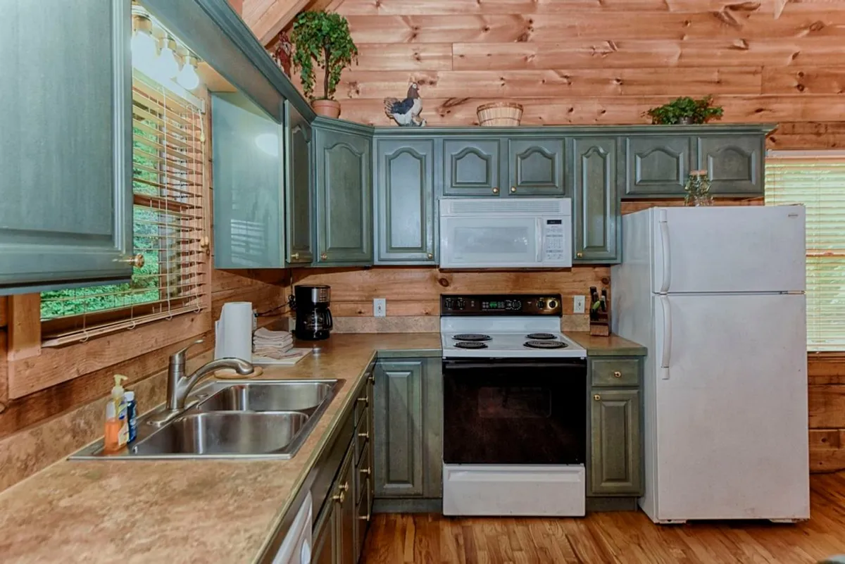 A view of the fully equipped kitchen featuring green cabinetry, a full stove, spacious countertops, a refrigerator, a deep farmhouse sink, and wood paneling that exudes a warm, homely feel. The setup clearly prioritizes functionality for meal preparation and group gatherings.