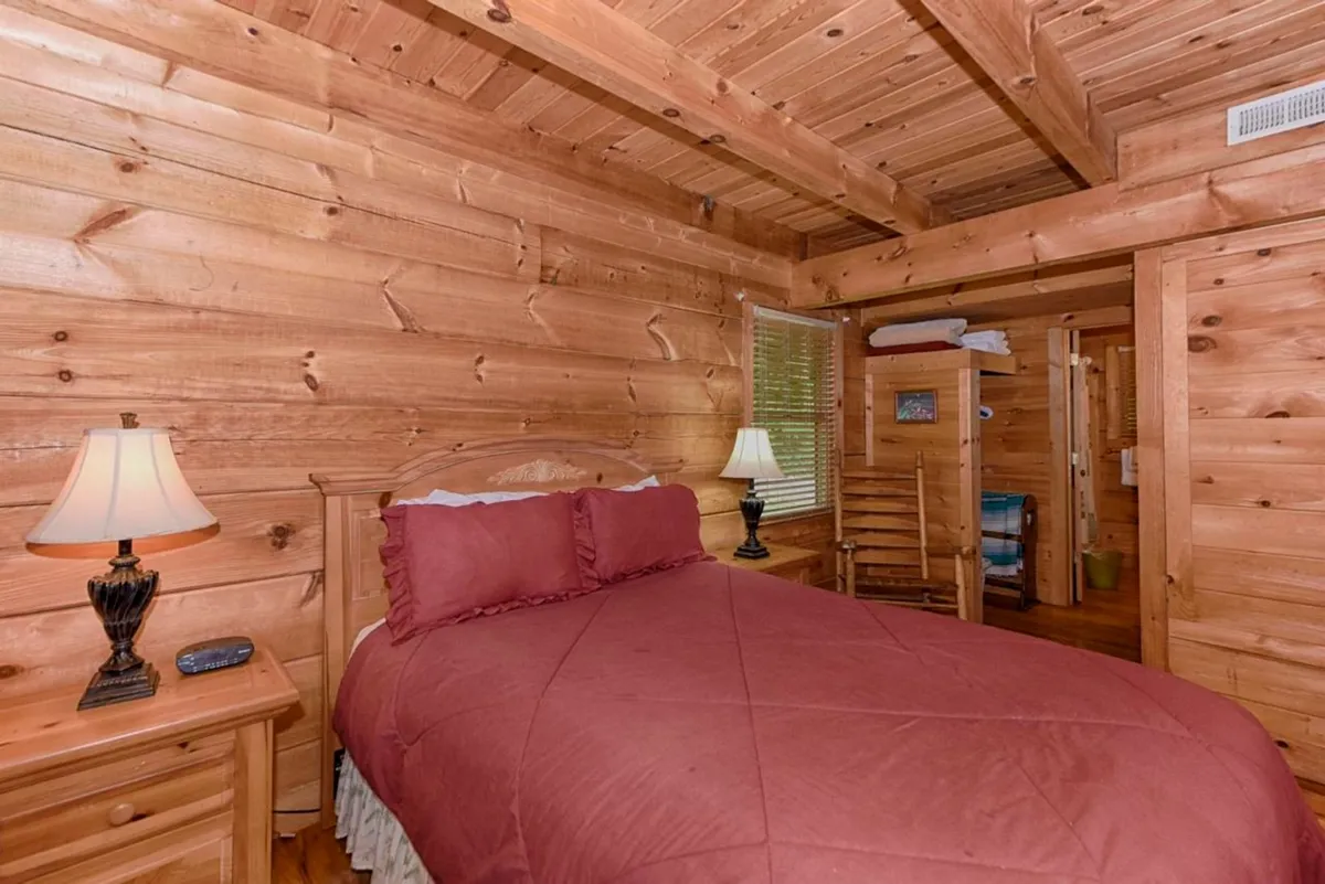 A cozy bedroom featuring a wooden bed, nightstand with lamp, rustic wooden walls, a window with blinds letting in natural light, and a wooden chair. The arrangement exudes warmth and comfort, ideal for restful nights.