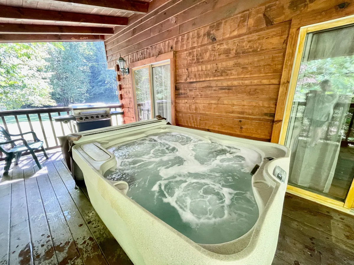 A view from the wooden deck showcases a hot tub, grill, and large windows, with the outdoors visible beyond. The deck opens to scenic views and plenty of space, seamlessly connecting the comfort of the home with the surrounding nature.