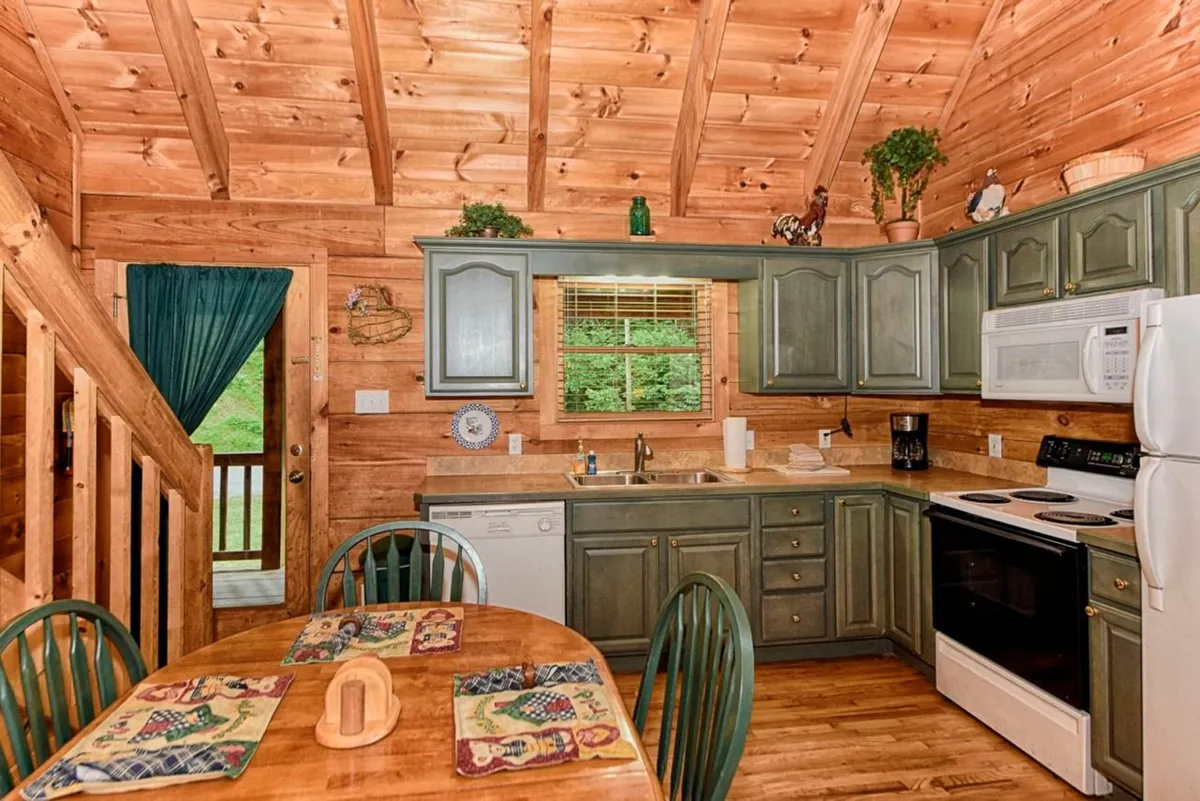 This image captures an inviting kitchen and dining area with wooden cabinets, a large farmhouse-style dining table, visible modern kitchen appliances such as a coffee maker and microwave, and a window offering a glimpse of the wooded surroundings. The warm wood tones and cozy layout evoke a sense of rustic charm paired with practical convenience.