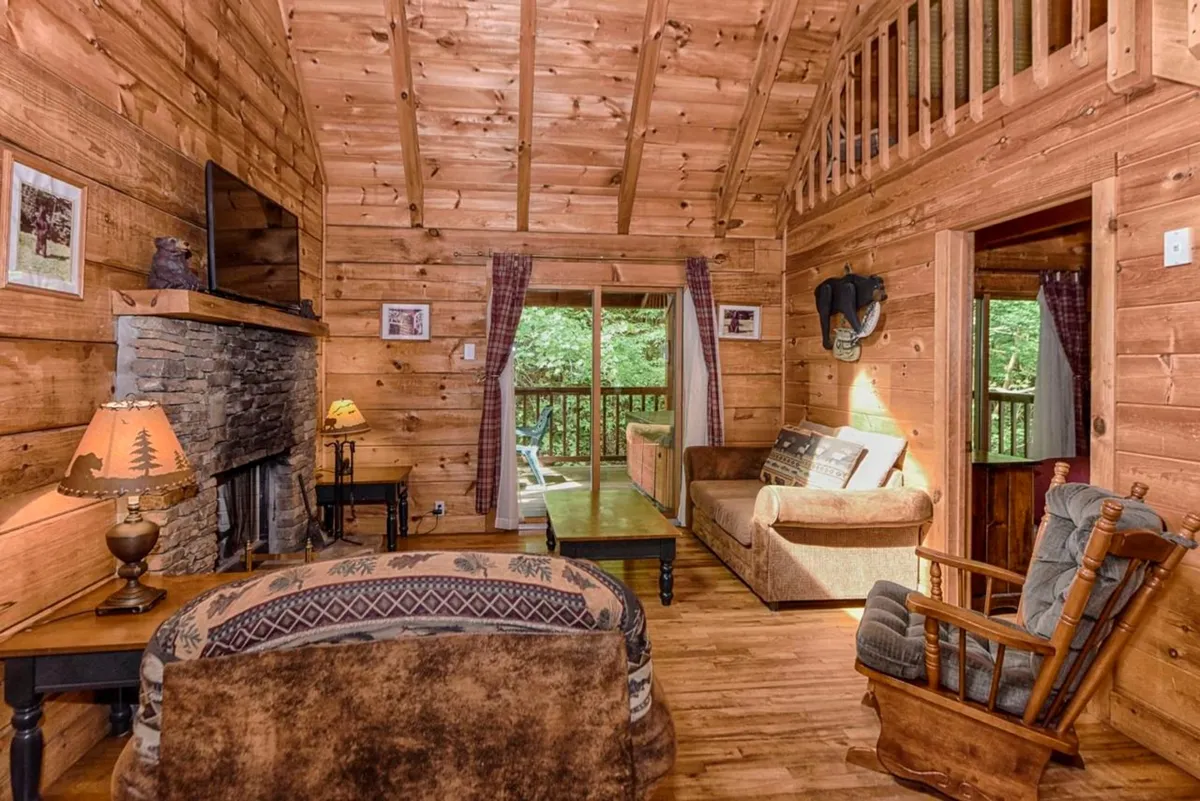 A cozy living room with a striking stone fireplace, sturdy wooden beams on the ceiling, large windows framing lush views of the outdoors, and comfortable seating arranged for conversation or relaxation. The natural light streaming in enhances the warm wood décor, creating a welcoming gathering space.
