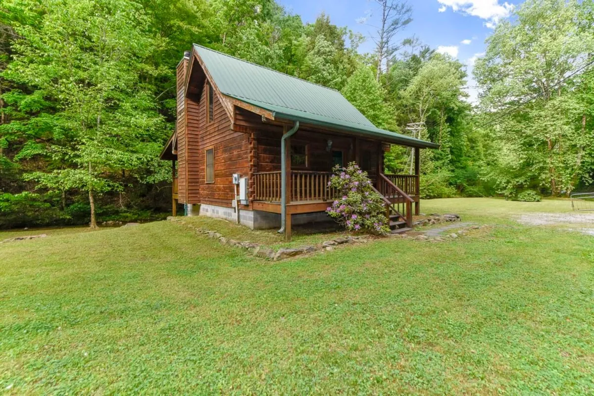 A classic log cabin with a green roof and welcoming front porch sits amidst blooming flowers and lush greenery. The porch offers an inviting spot to relax, enjoy morning coffee, or socialize while surrounded by nature.