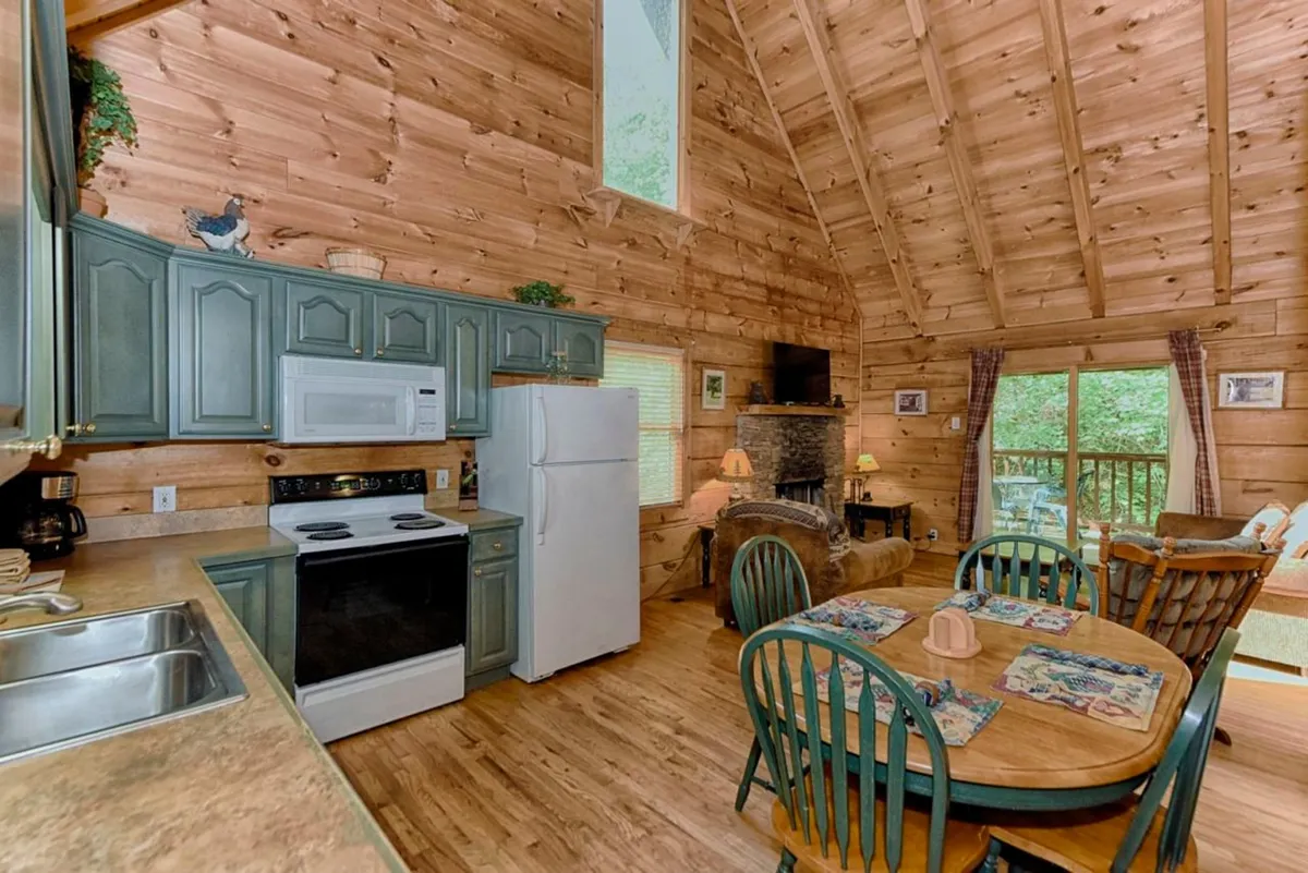 An open-plan interior featuring a dining table set near a rustic fireplace, modern kitchen appliances, and large windows that allow plenty of natural light. The scene evokes a space designed for socializing, dining, and relaxing, emphasizing a seamless blend between the living and dining areas.